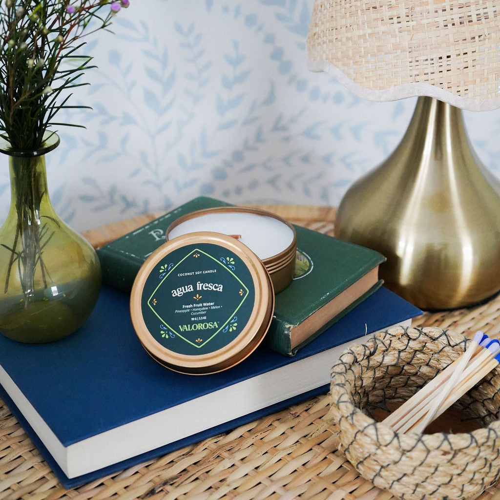 Agua Fresca Candle Tin on a wicker side table with a lamp, matches and lavender floral arrangement on top of two books