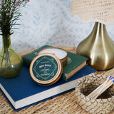 Agua Fresca Candle Tin on a wicker side table with a lamp, matches and lavender floral arrangement on top of two books