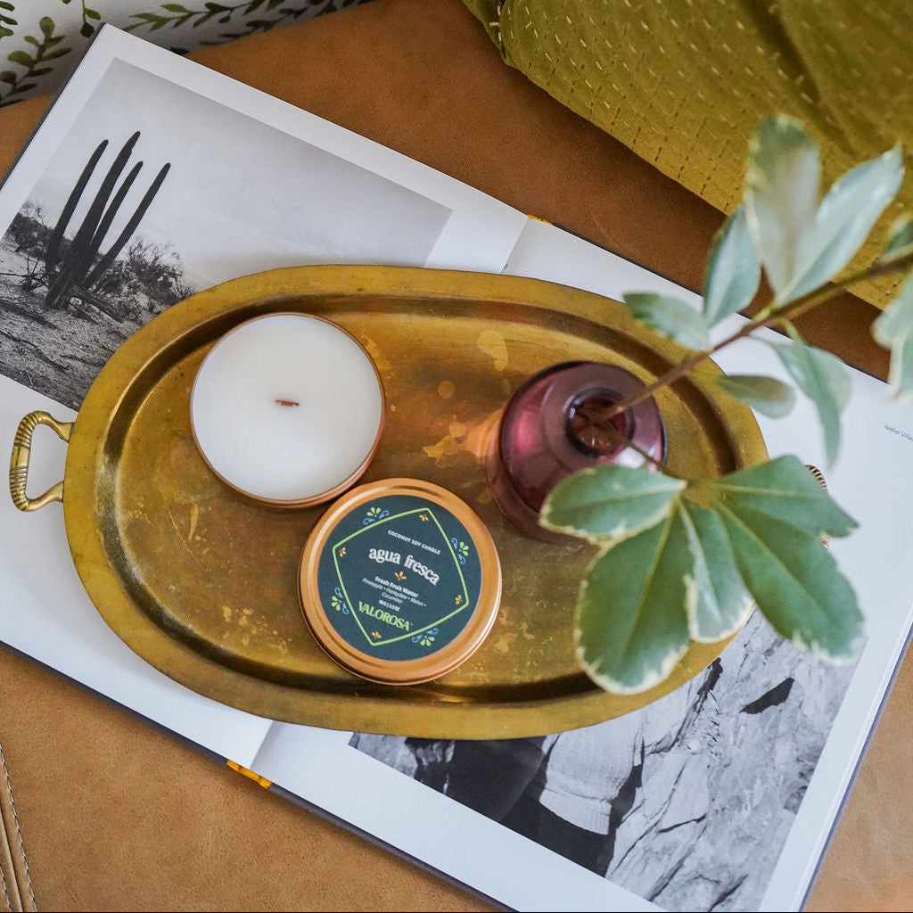 Agua Fresca Candle Tin looking overhead on a gold tray on top of an open book with a purple vase and greenery
