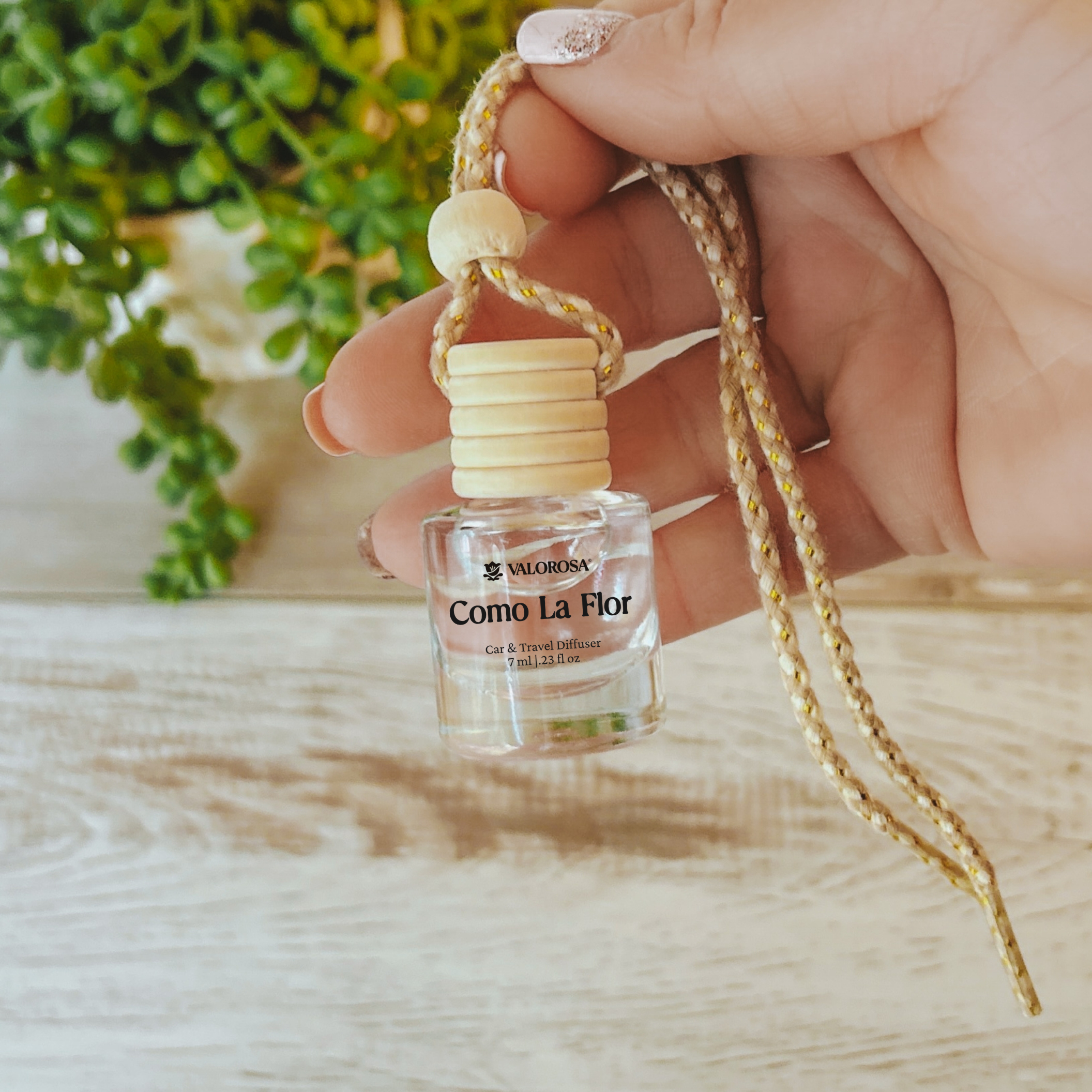 Small car diffuser bottle with a string held by a hand on a wooden surface with a blurred plant in the background