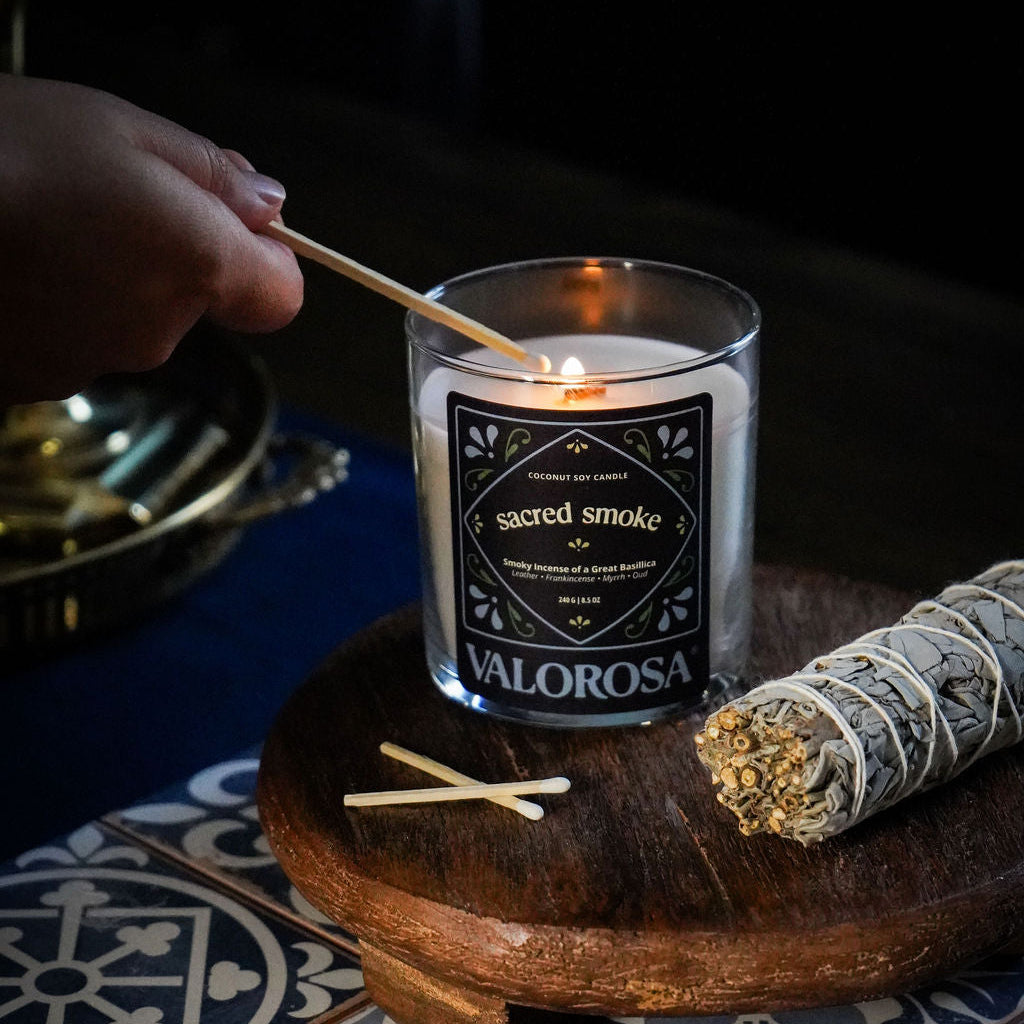 Valorosa Sacred Smoke Candle being lit on a wooden tray with a rolled sage on a patterned tile floor.