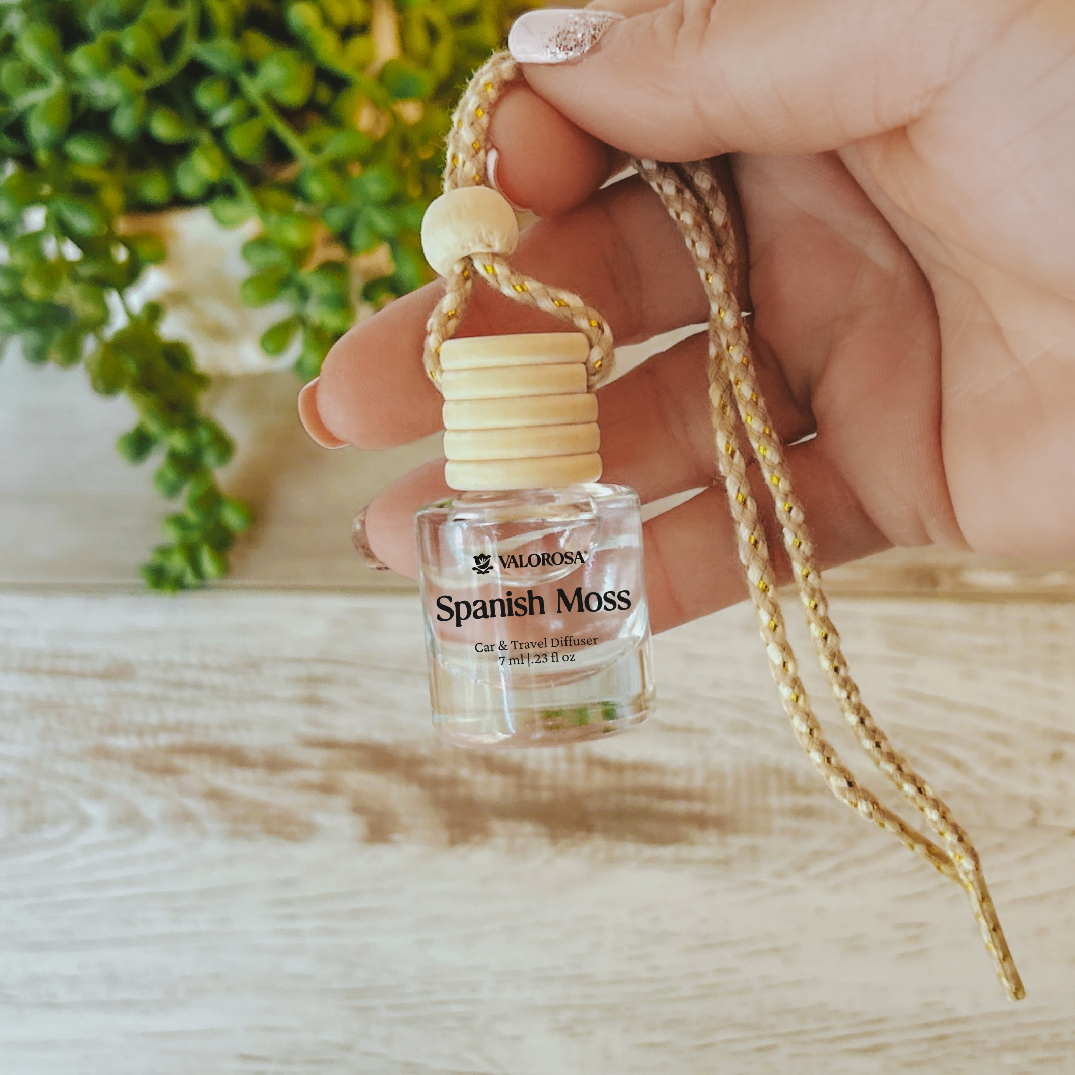 Small car diffuser bottle with a string held by a hand on a wooden surface with a blurred plant in the background