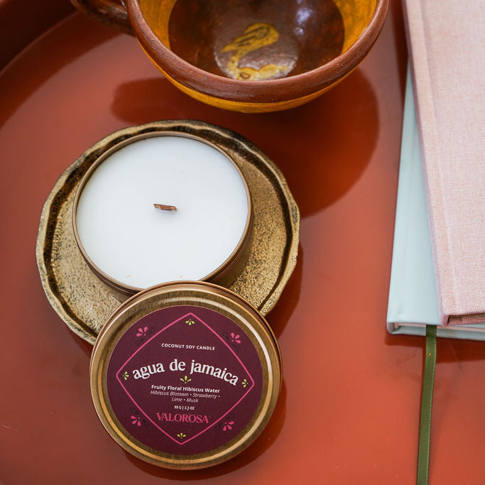 Candle in a decorative tin with 'agua de Jamaica' label on a red surface with a notebook and pen.