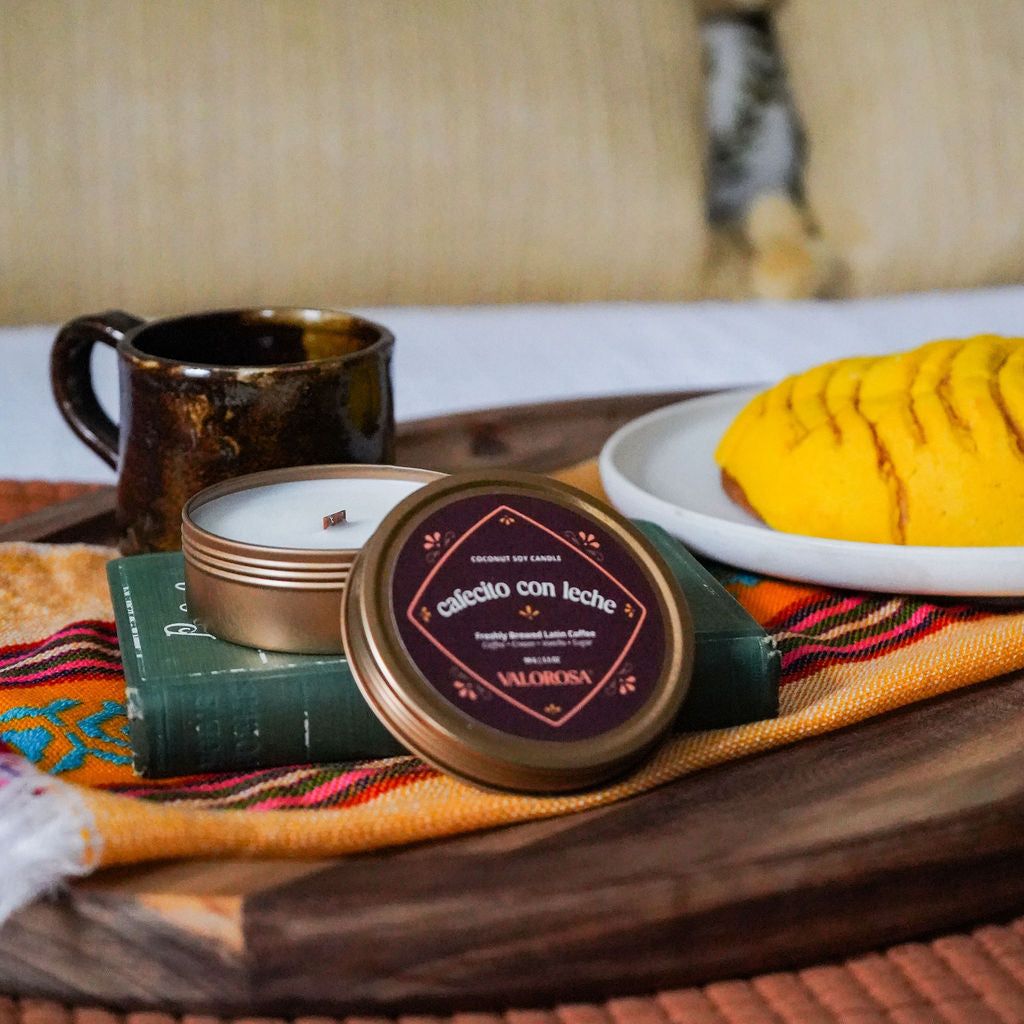 Cafecito Con Leche Candle Tin, mug, and plate with food on a wooden tray with a colorful cloth, against a blurred indoor background.