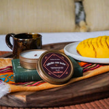 Cafecito Con Leche Candle Tin, mug, and plate with food on a wooden tray with a colorful cloth, against a blurred indoor background.