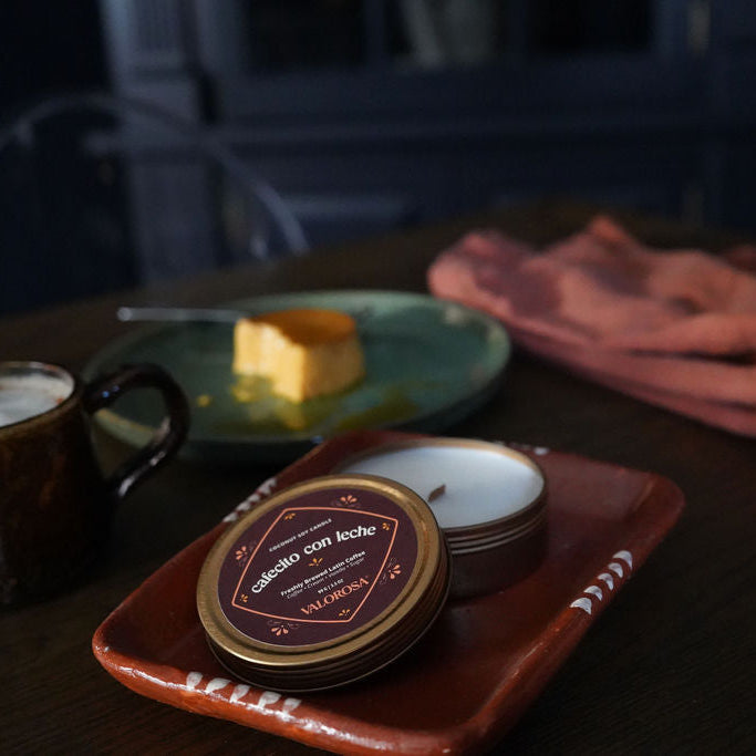 Cafécito con leche candle tin in a brown cup on a wooden table with a tray of pastries and a teapot.