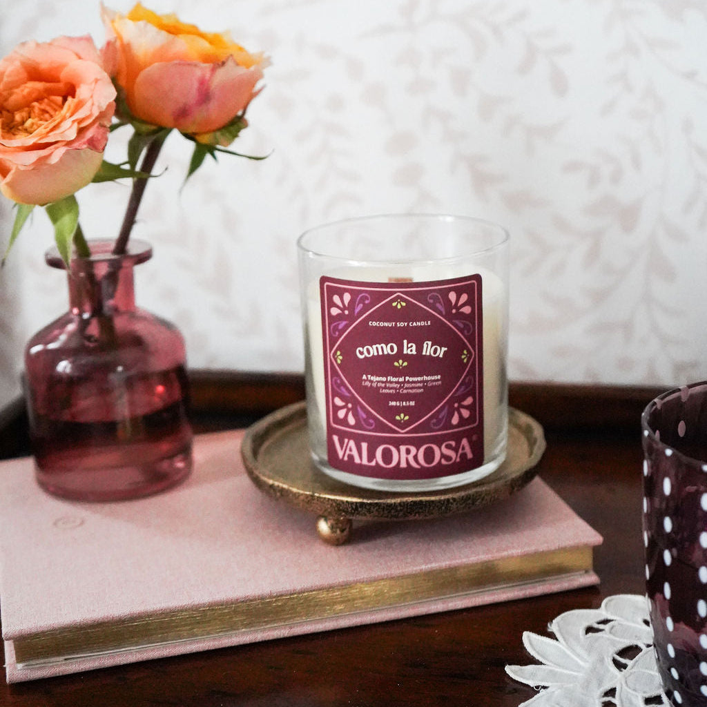 Valorosa Como La Flor Candle on a wooden surface with a vase of flowers and a book in the background.