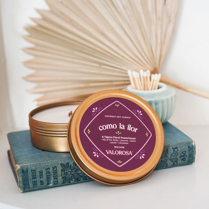 Como La Flor Candle with a purple label on a book, surrounded by dried plants on a white surface