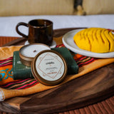 Coquito Candle with decorative packaging on a wooden tray with a plate of food and mug in the background.