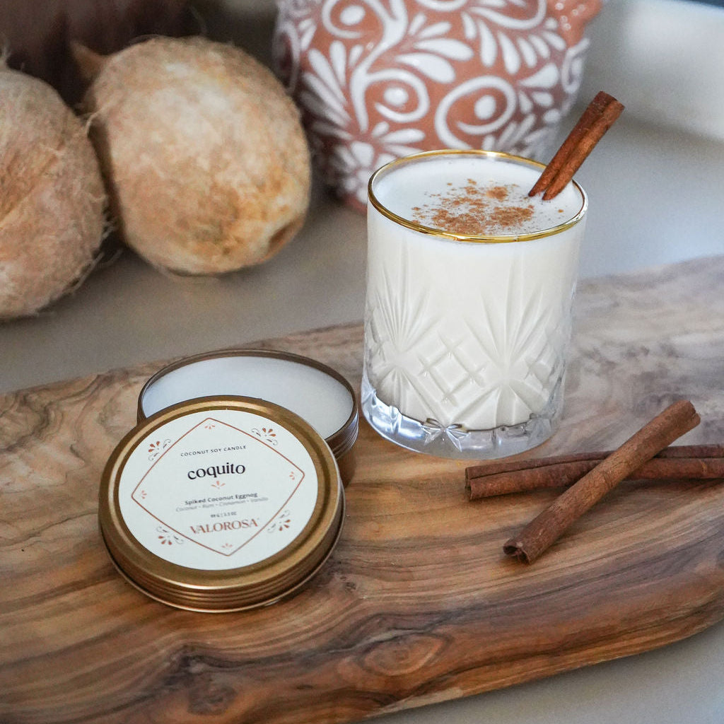 Glass of coquito with cinnamon stick on a wooden tray, coconut, decorative vase and a coquito candle tin in the background.