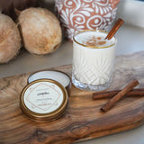 Glass of coquito with cinnamon stick on a wooden tray, coconut, decorative vase and a coquito candle tin in the background.