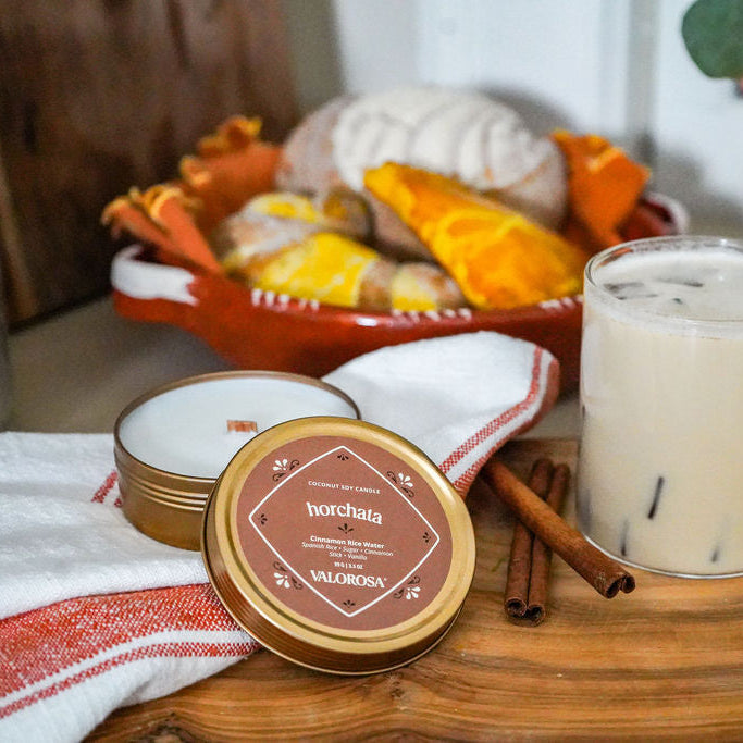 Glass of horchata with a straw on a wooden cutting board, surrounded by an horchata candle tin and decorative items.