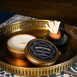 Sacred Smoke candle tin, incense stick holder, and on a decorative tray with a patterned surface.