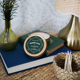 Candle tin with packaging on books next to a vase with flowers and a lamp.