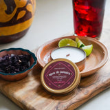 A candle of 'agua de Jamaica' on a wooden board with a candle and limes, next to a glass of red liquid.