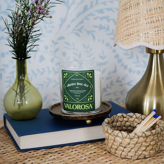 Candle labeled 'Valorosa' on a book with a vase, lamp, and basket on a textured surface.