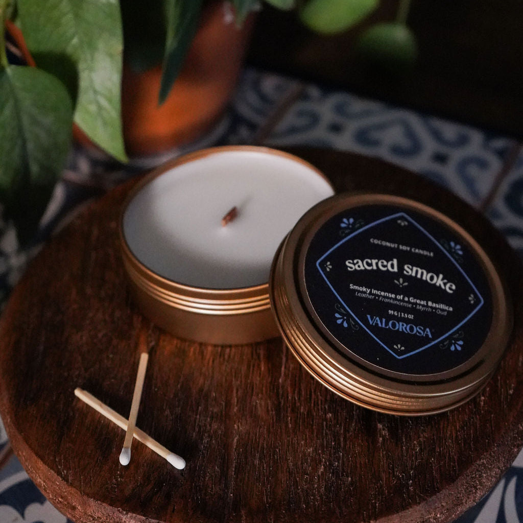Sacred Smoke Candle tin and packaging on a wooden surface with plants in the background